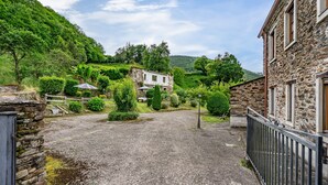 Paisagem rural com um edifício de pedra, entrada de cascalho e vegetação exuberante.