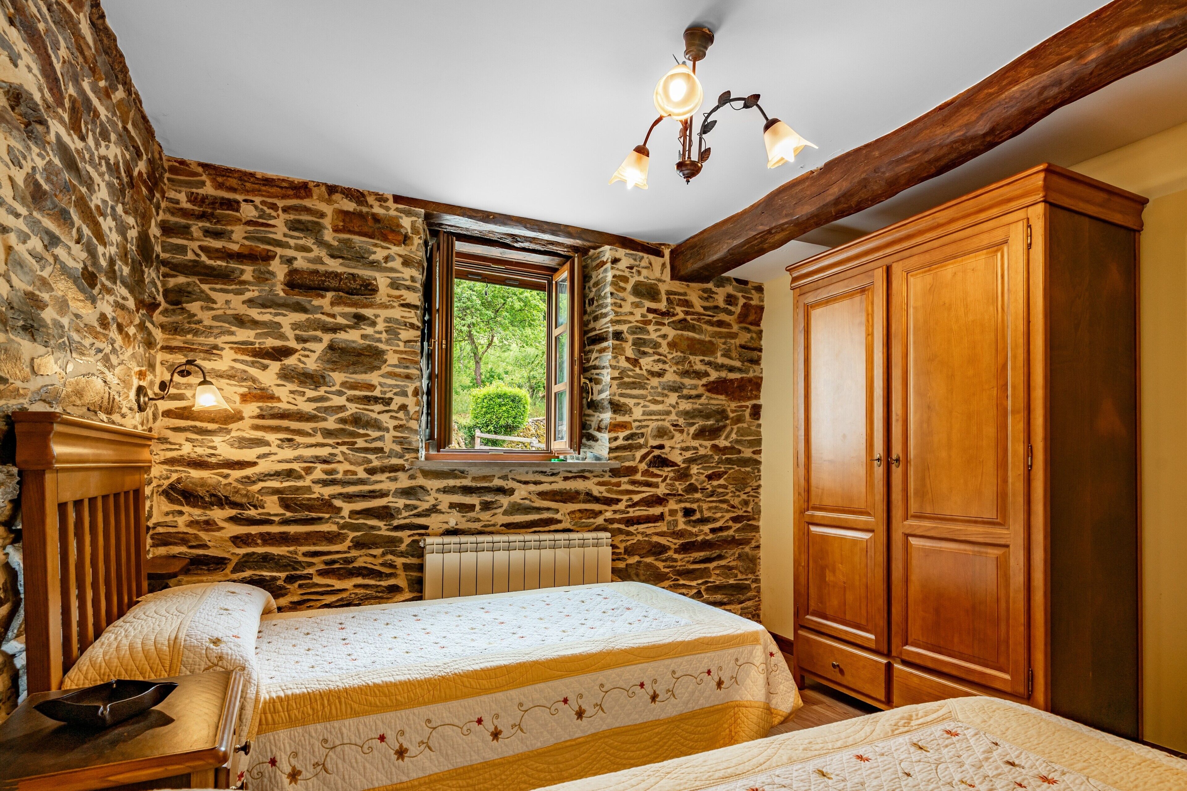 Quarto com parede de pedra, cama de madeira, guarda-roupa e janela com vista para o verde.