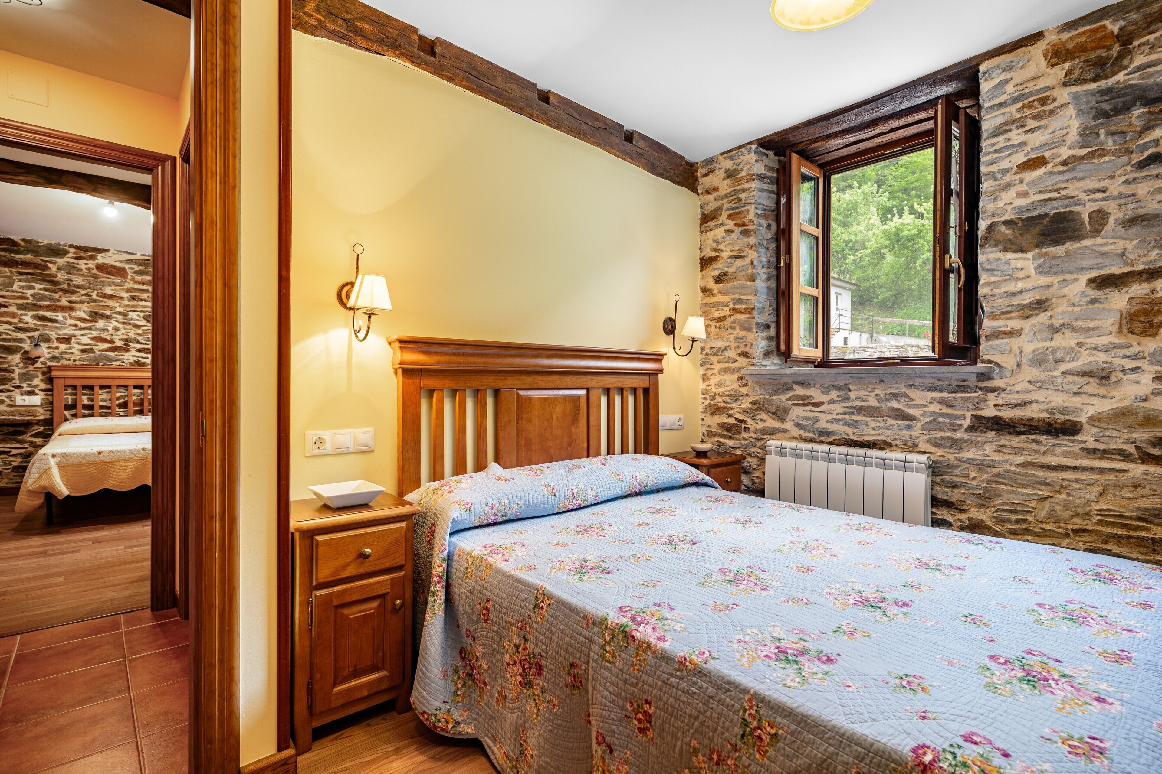 A bedroom with a wooden headboard, a floral bedspread, a stone wall, and a window with a view.