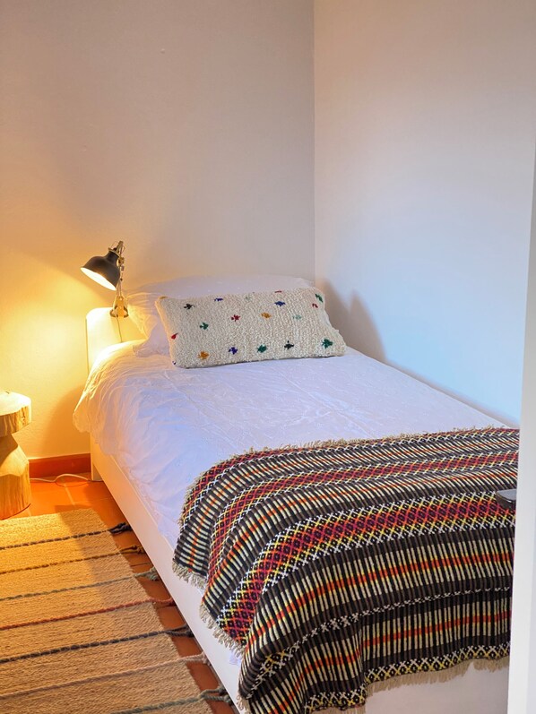 Una cama bien hecha con un edredón blanco y una manta colorida, una lámpara de noche y un pequeño taburete de madera.