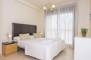 Quarto com uma cama grande, mesas de cabeceira, um criado-mudo, um candelabro, uma janela com cortinas finas e uma pequena planta em vaso.