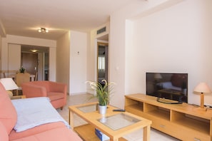 Uma sala de estar moderna com um sofá rosa, uma mesa de centro de vidro, uma TV sobre um suporte de madeira e uma planta em vaso.