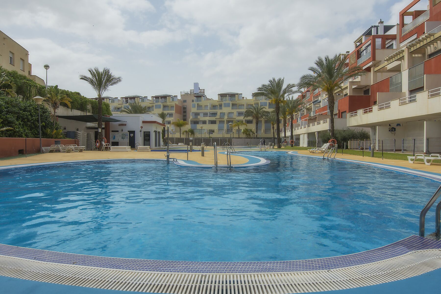 A swimming pool with a tiled edge, surrounded by palm trees and a building with balconies.