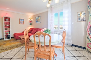 Un salon chaleureux avec un canapé rouge, une table à manger ronde entourée de chaises, une bibliothèque et une grande fenêtre agrémentée de rideaux.