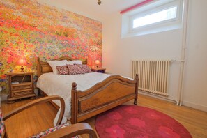 Une chambre à coucher avec un lit en bois, une tapisserie à motifs floraux, un tapis rouge, une table de chevet et une fenêtre donnant sur un beau paysage.