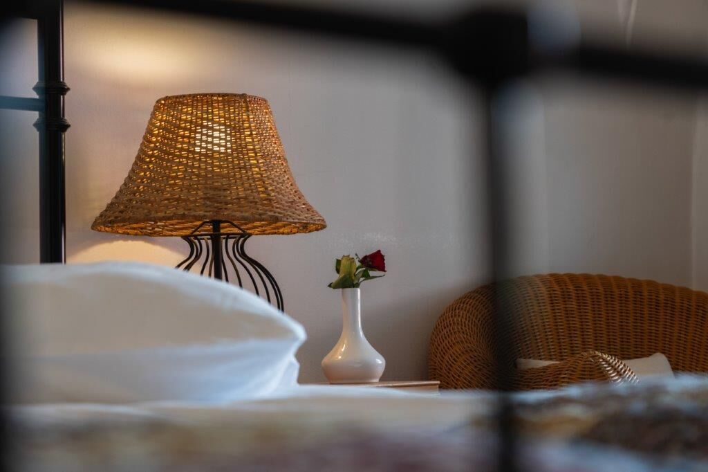 A bed with white bedding, a wicker lamp, and a white vase with a single rose.
