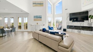 A spacious living room with a large sofa, a TV mounted on a marble fireplace, and a balcony with a dining area. - Living Luxe - Oceanfront holiday bliss! (St. Augustine Beach)
