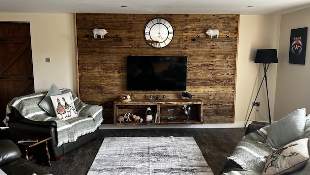 A living room with a wooden wall, a mounted clock, a flat-screen TV, and a console table with decorative items. - Luxury Country Lodge in Bretherton -sleeps 1 Double bed & 1 bed Settee (Leyland)
