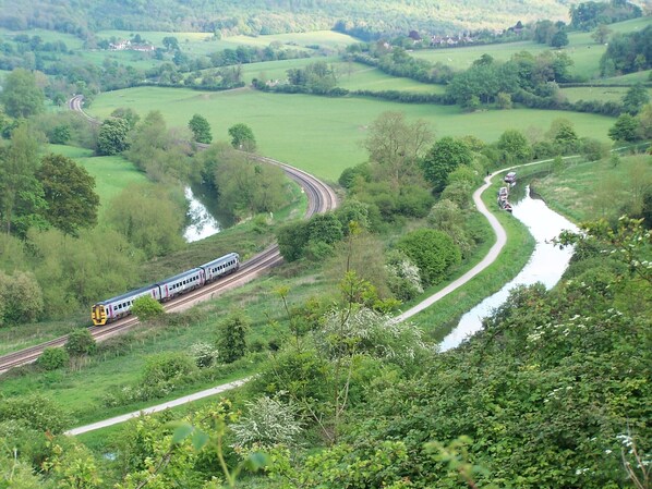 A train traveling through a lush green valley with a winding road and a river. - The Cru Houseboats (Bath)