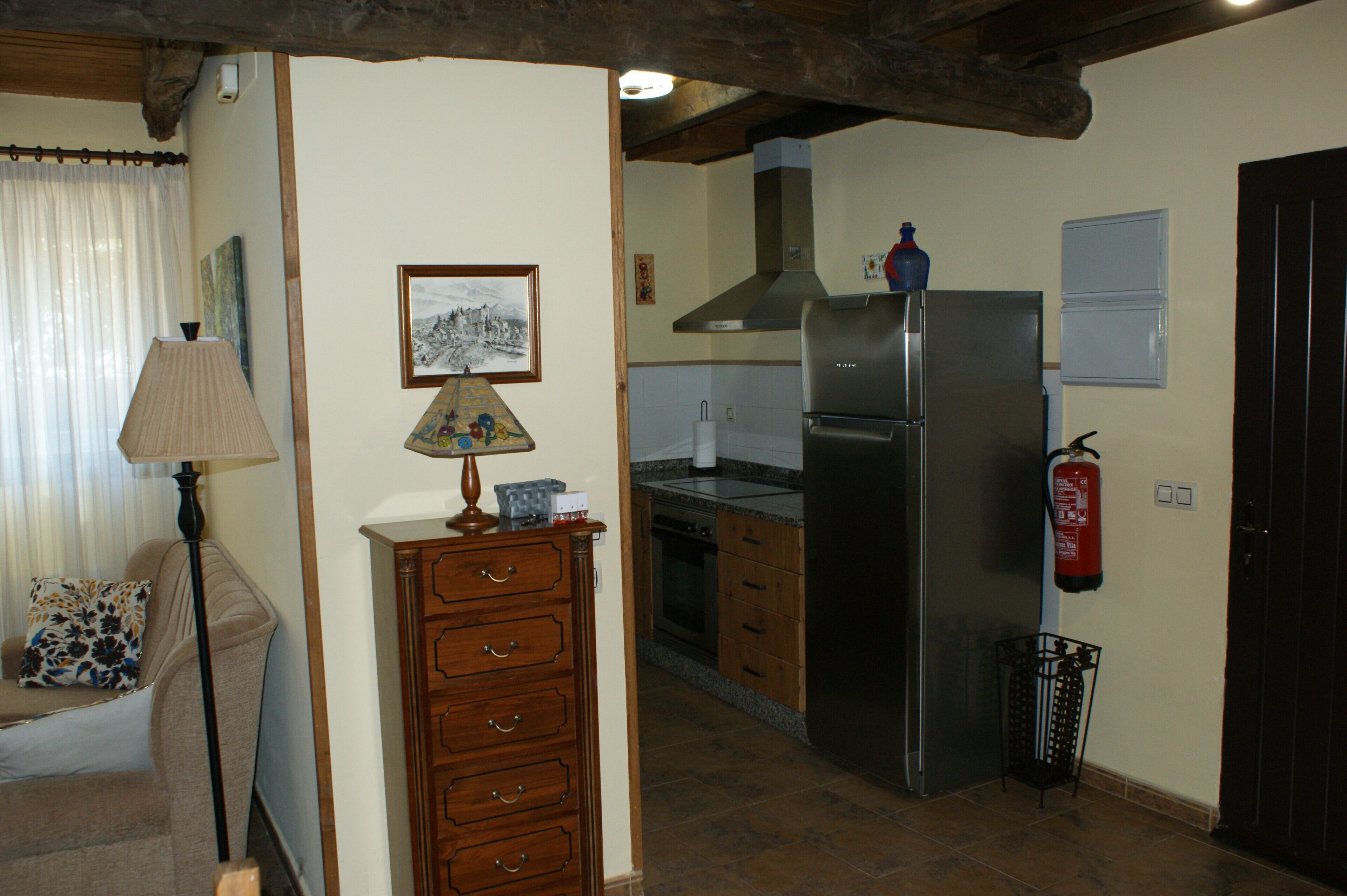 A kitchen with wooden cabinets, a stainless steel refrigerator, a fire extinguisher, and a dining area with a lamp and a sofa.