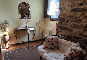 A cozy room with a wooden floor, a white sofa with floral cushions, a small table with a vase of flowers, a mirror, a radiator, and a brick wall.