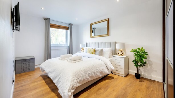 Quarto com cama grande, mesa de cabeceira, luminária, espelho, planta em vaso e cesto de roupa.