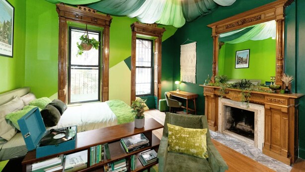 Une pièce avec un mur vert, une cheminée en bois, un lit recouvert d’une couverture verte, un bureau avec une lampe et une bibliothèque.