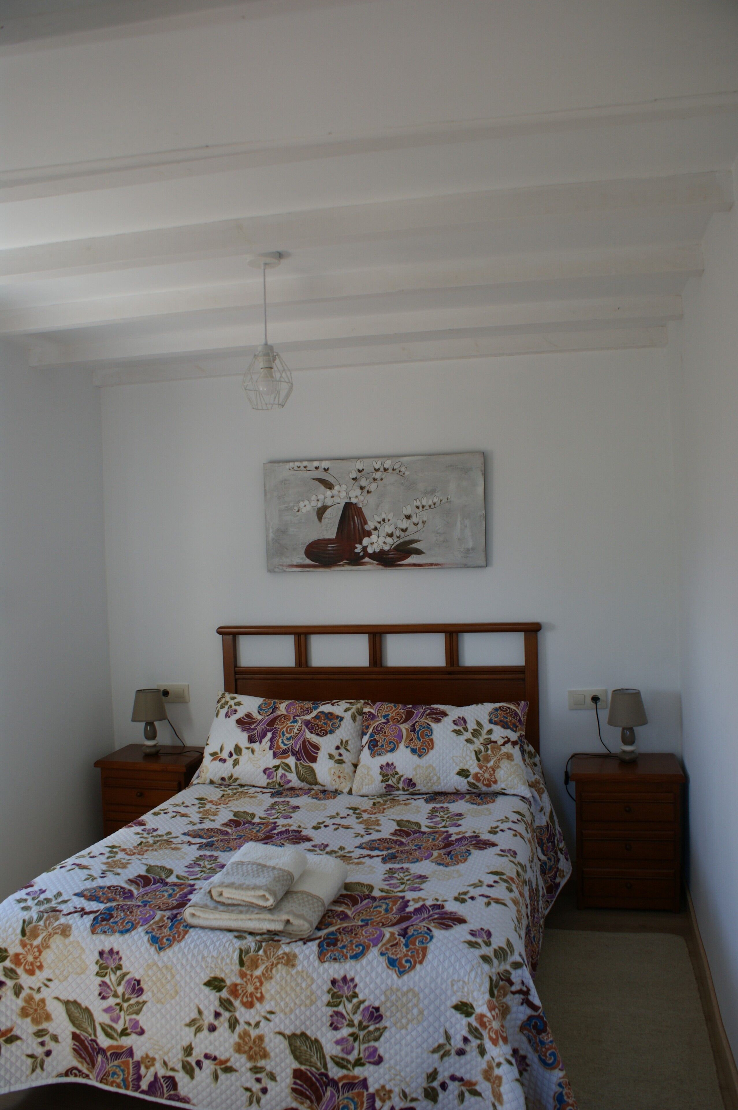 Un dormitorio con cabecera de madera, ropa de cama floral, dos mesitas de noche con lámparas y una obra de arte colgada en la pared.