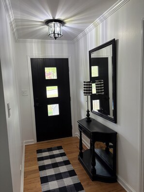 Un couloir avec une porte noire, un miroir, une console noire et un tapis à carreaux noirs et blancs.