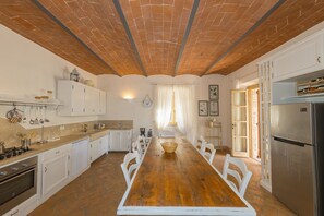 Een keuken met een houten tafel, witte kasten, een koelkast en een plafond van baksteen.