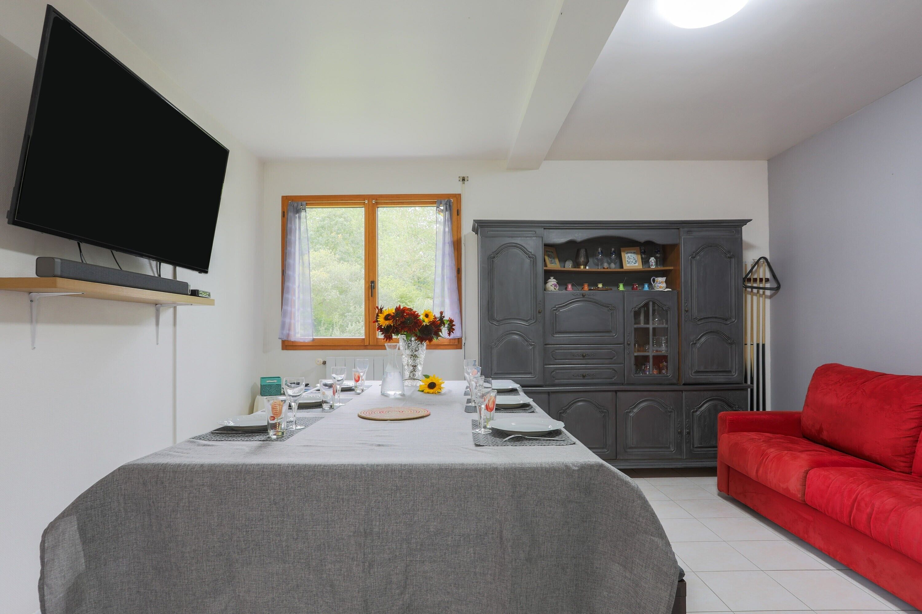 Ein Essbereich mit einem Tisch für vier Personen, einem an der Wand montierten Fernseher und einem roten Sofa im Hintergrund.