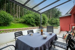 A covered patio with a table set for dining, surrounded by chairs, and a view of a forested area. - Annexe du Petit Echo de la mode – Nature Gîte with Private Garden, Billiards, Wi-Fi, Wood Stove (Plélo)