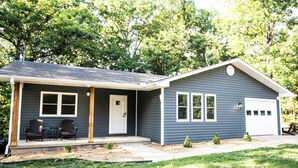 A single-story house with a front porch, two windows, a garage, and a small front yard with a lawn and a few shrubs. - The Ridge House-Lake House w/Hot Tub & Game Room-DOG friendly (Eureka Springs)