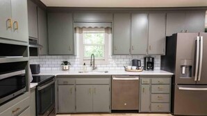 A modern kitchen with grey cabinets, stainless steel appliances, a window with a white curtain, and a wooden floor. - The Ridge House-Lake House w/Hot Tub & Game Room-DOG friendly (Eureka Springs)