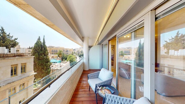A balcony with a wooden floor, two cushioned chairs, a small table with magazines, and a glass door leading to a room. - King David Residences: Spacious 3-bdrs 3-bths with Pool, Gym & View in Jerusalem (Jerusalem)