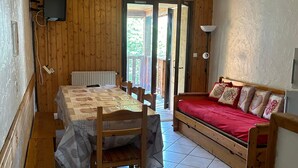 A room with a wooden table, chairs, a bed with red bedding, a television, and a door leading outside. - Apartment 'La Sétaz' with Mountain View, Private Terrace and Wi-Fi (Valloire)
