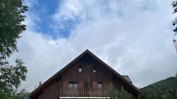 A two-story wooden chalet with a balcony on the upper floor, surrounded by trees and a mountainous backdrop.