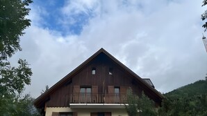 A two-story wooden chalet with a balcony on the upper floor, surrounded by trees and a mountainous backdrop.