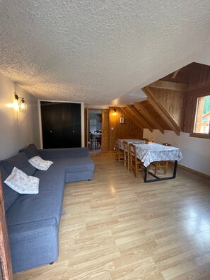 A room with a sectional sofa, a dining table with chairs, wooden beams on the ceiling, and a view through a window. - Apartment 'Le Galibier' with Mountain View, Shared Garden and Wi-Fi (Valloire)