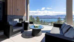 Ein moderner Außenbereich mit Korbmöbeln, einem runden Tisch mit floraler Dekoration und Blick auf einen See und Berge.