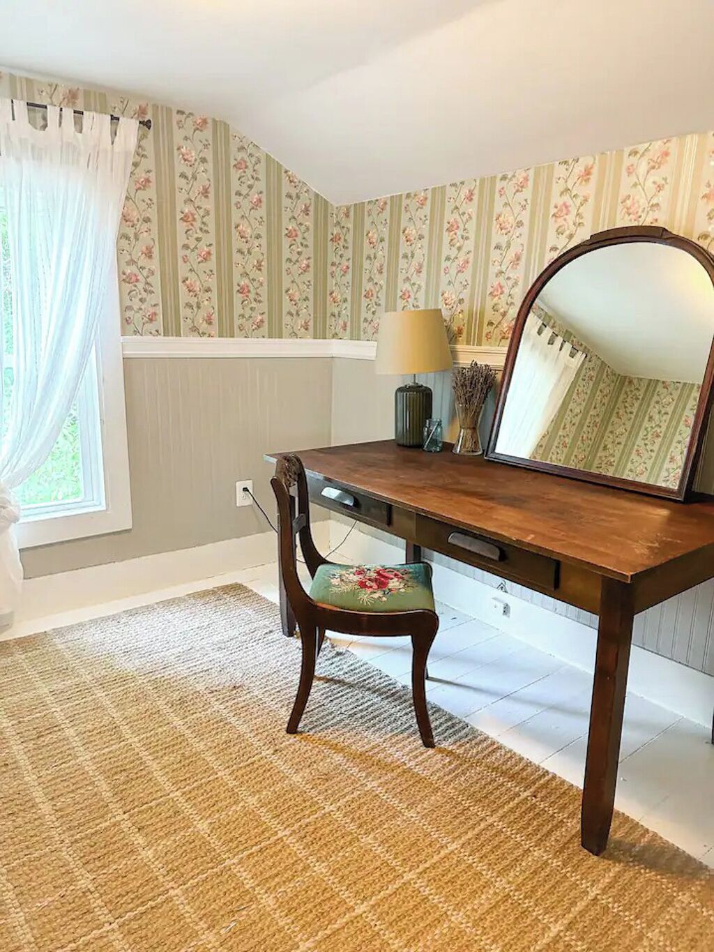 A dining room with a wooden table, a chair with a floral cushion, a lamp, and a mirror.