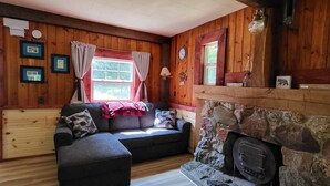 A cozy cabin interior with a stone fireplace, wooden walls, a sofa, and a window with curtains. - Green Cabin in the Woods (Irons)
