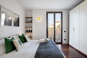 A bedroom with a bed, pillows, a nightstand, a shelf with decorative items, a door to a balcony, and a wall-mounted light.