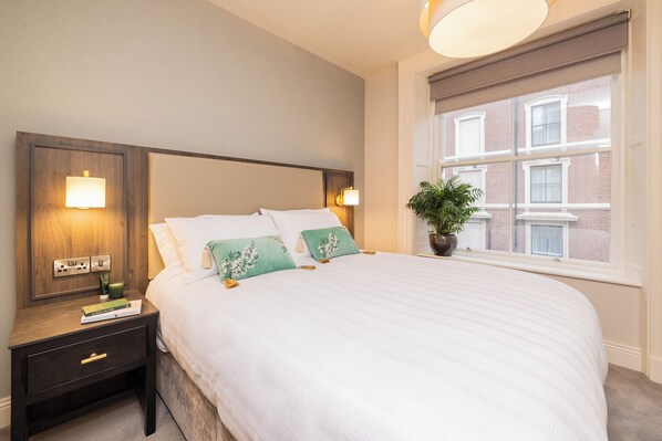 A neatly made bed with white bedding and green floral pillows, a bedside table with a lamp, and a window with a view of a brick building.