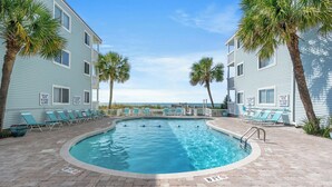 A swimming pool with lounge chairs and palm trees, surrounded by light blue buildings. - Ocean Garden Villas J1- 2BR/2BA- Pool (North Myrtle Beach)