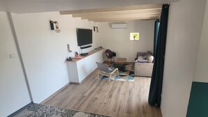 Een moderne leefruimte met een houten vloer, een flatscreen televisie aan een witte muur, een bureau met stoel en een bank met kussens.