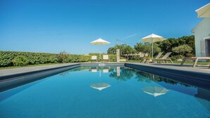 A swimming pool with lounge chairs and umbrellas, surrounded by greenery and a clear sky.