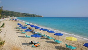 A beach with rows of sun loungers and umbrellas, a clear blue sea, and a hilly landscape in the background.