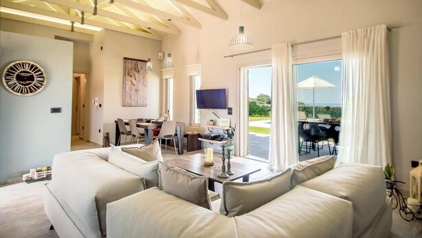 A spacious living area with a large sofa, a dining table, and a television. There is a balcony with outdoor furniture and a view of the landscape.
