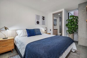 A modern bedroom with a large bed, bedside tables, a nightstand, a potted plant, and a bathroom visible through an open door. - Park38 (Envigado)