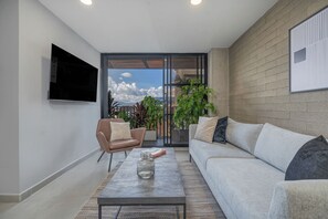 A modern living room with a sofa, armchair, and coffee table. There is a TV mounted on the wall and a large window with a view of the city. - Park38 (Envigado)