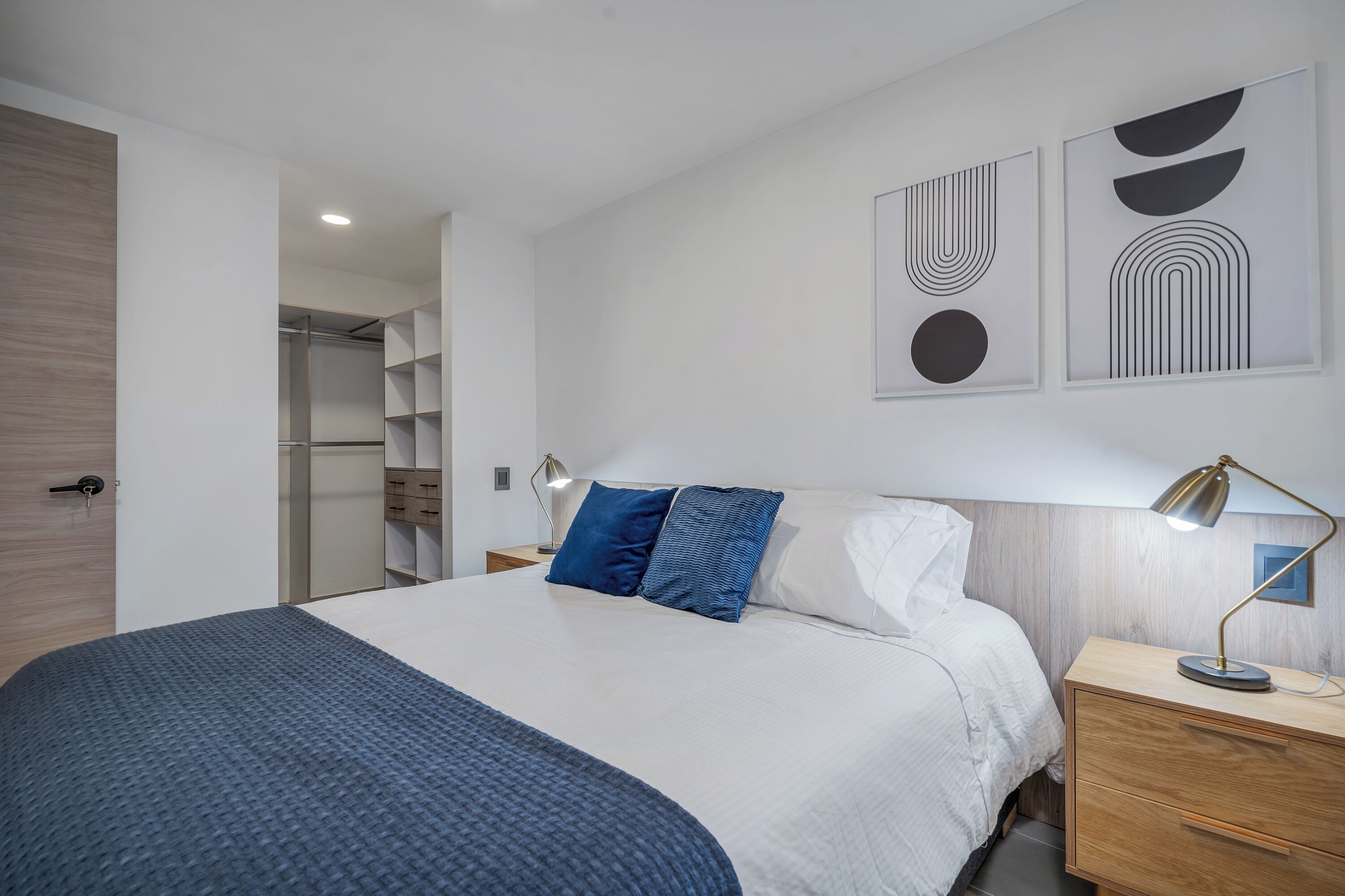 Une chambre à coucher moderne avec un grand lit, une table de chevet, une lampe de bureau et des œuvres d’art abstraites accrochées au mur.