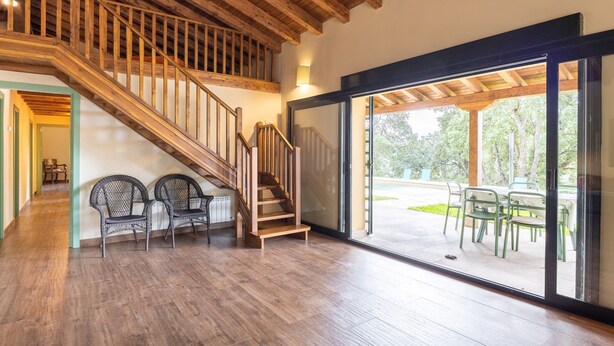 A spacious room with a wooden staircase, a sliding glass door leading to an outdoor patio, and a seating area with chairs.