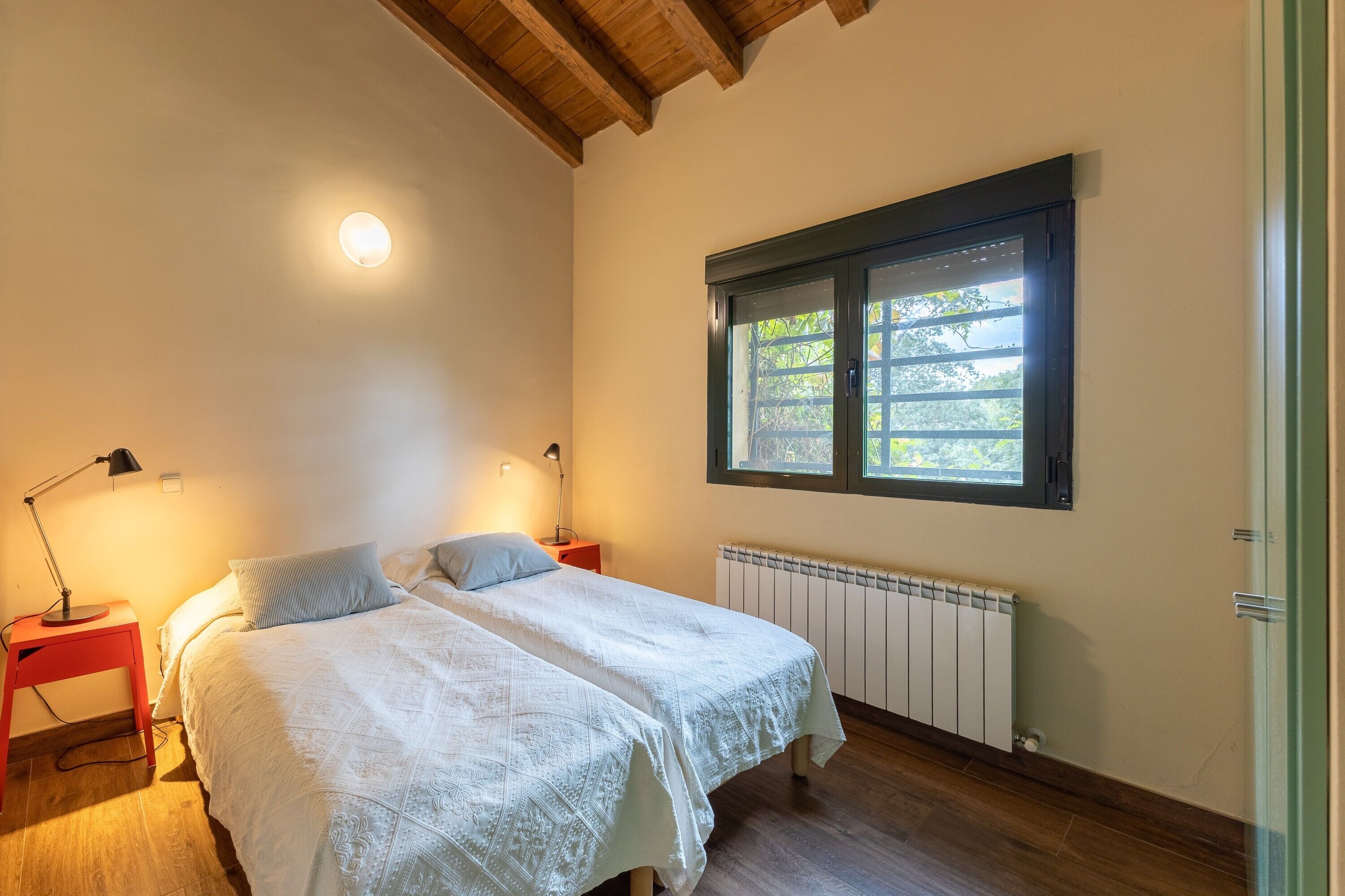 A bedroom with a double bed, two bedside lamps, a window with a view, a radiator, and a wooden ceiling.