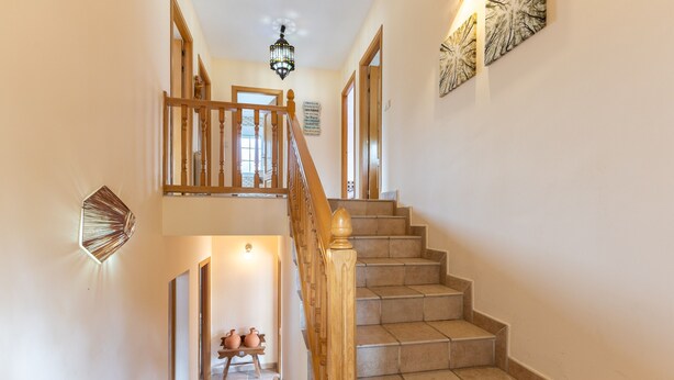 Un escalier avec des rampes en bois et des marches de couleur beige, menant à un palier agrémenté d’un lustre et d’œuvres d’art murales.