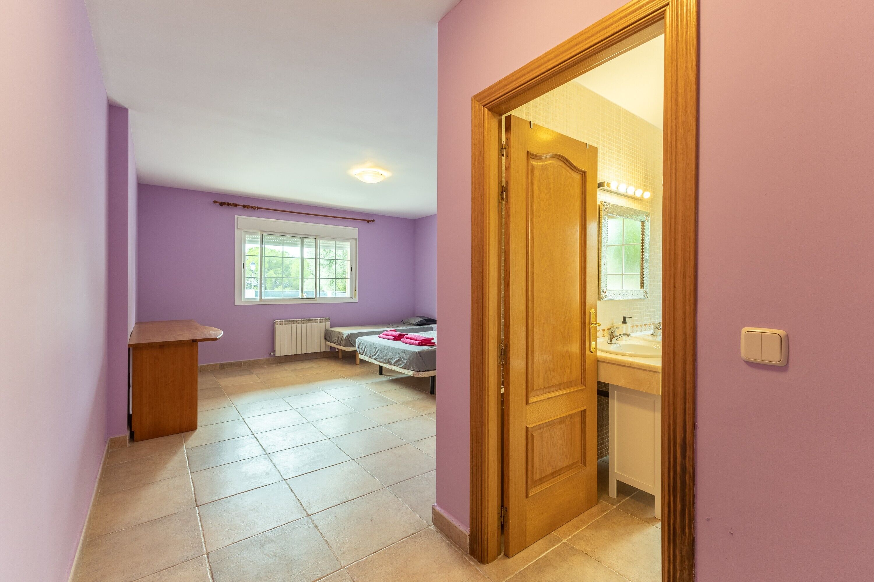 Een kamer met een bed, een houten bureau, een raam en een badkamer met een spiegel en wastafel.