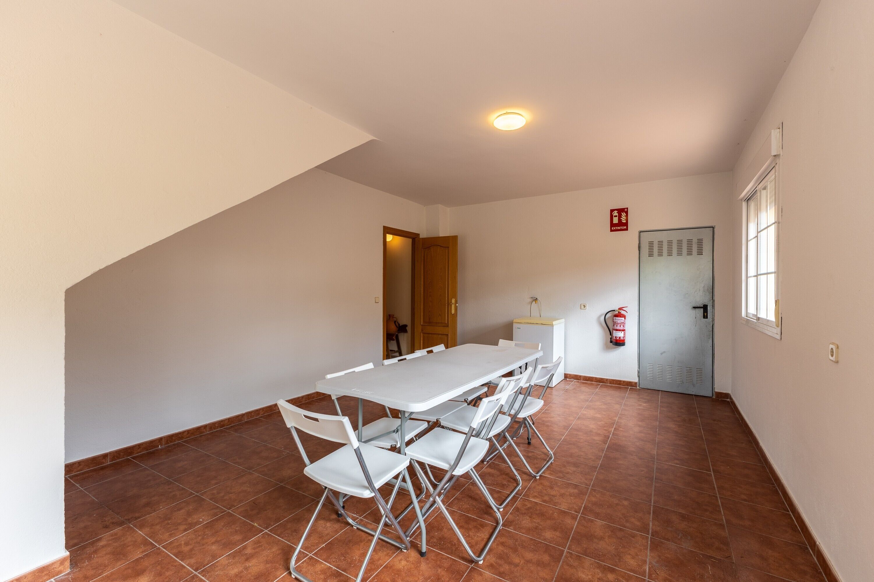 Una habitación con una mesa y sillas blancas, una puerta, un extintor y una ventana.