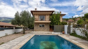 Une piscine bordée de pierres, une maison à deux étages dotée d’un balcon et un paysage montagneux en arrière-plan.