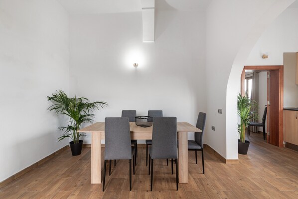 Un comedor con una mesa y sillas de madera, dos plantas en macetas y una lámpara en la pared.