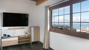 A room with a TV mounted on the wall, a wooden cabinet, a window with a view of a coastal landscape, and a ceiling with exposed beams.